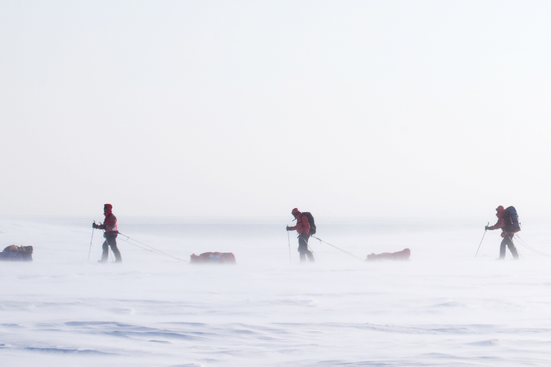 Three figures in red jackets and backpacks trek across a vast, windy, snow-covered plain, each pulling a sled.