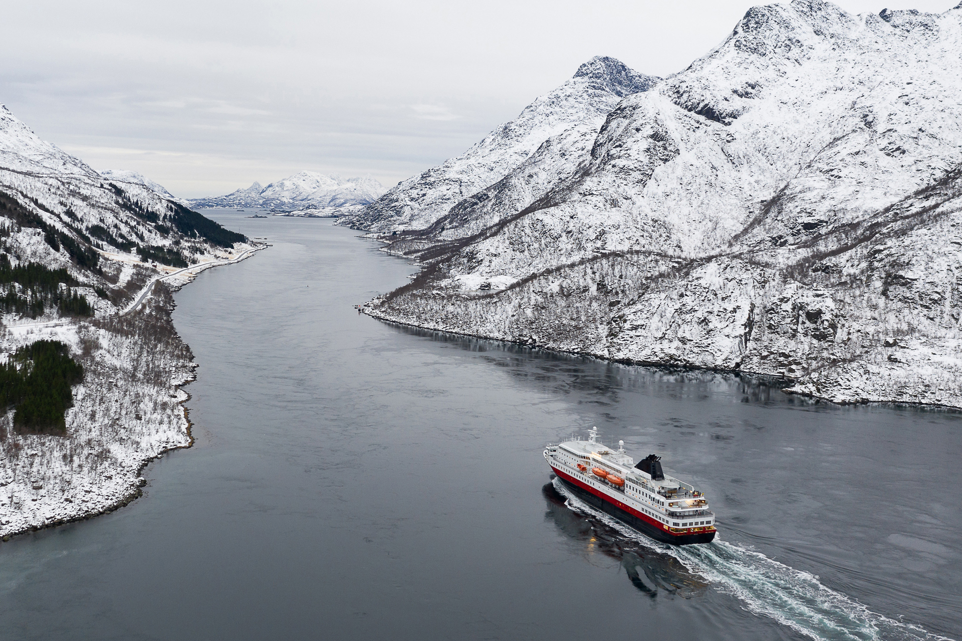 Un crucero rojo y blanco recorre una oscura vía navegable rodeada de montañas nevadas bajo un cielo nublado.