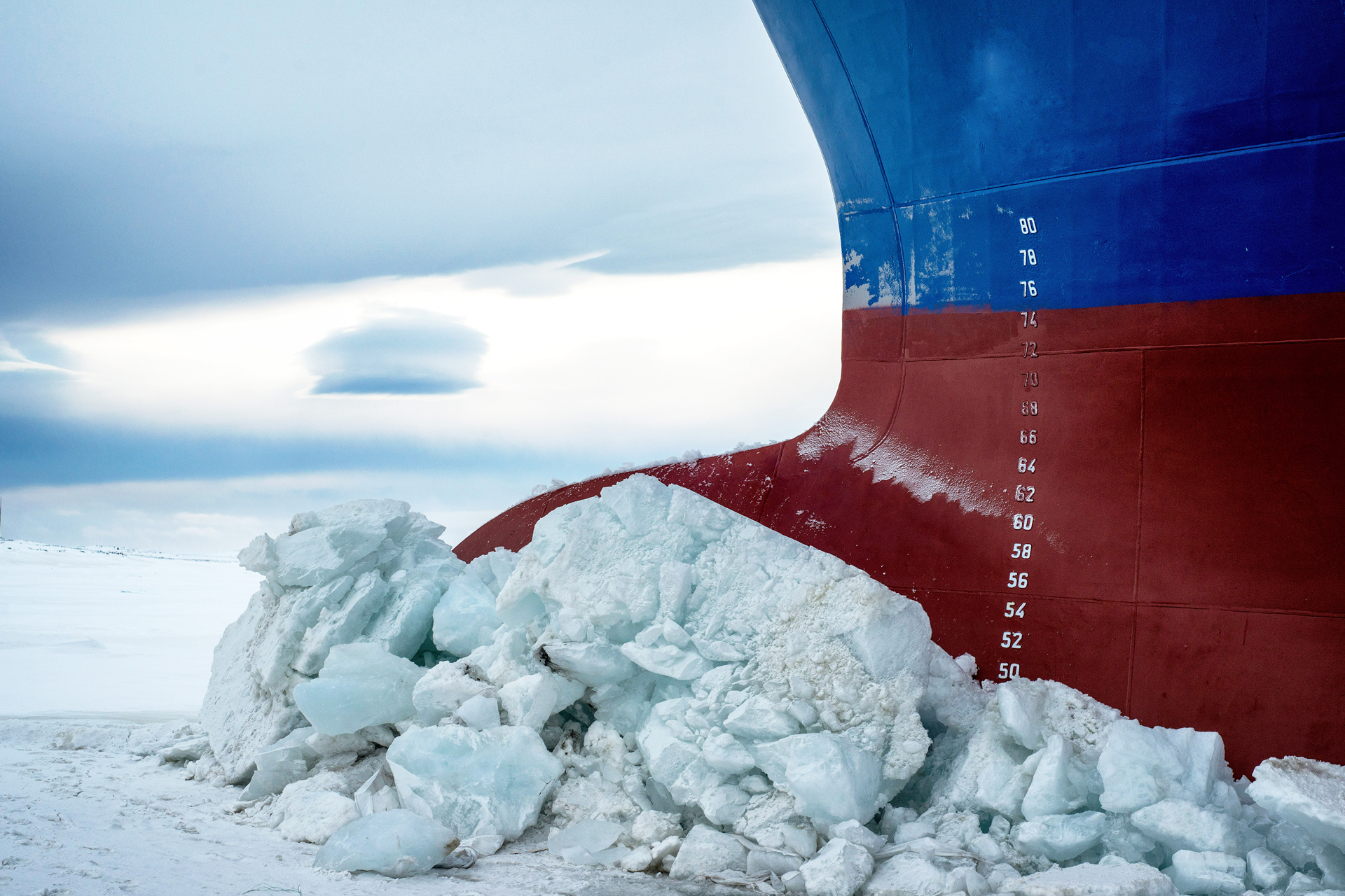 The red and blue hull of a large ship is surrounded by broken chunks of white ice and snow under a cloudy sky.
