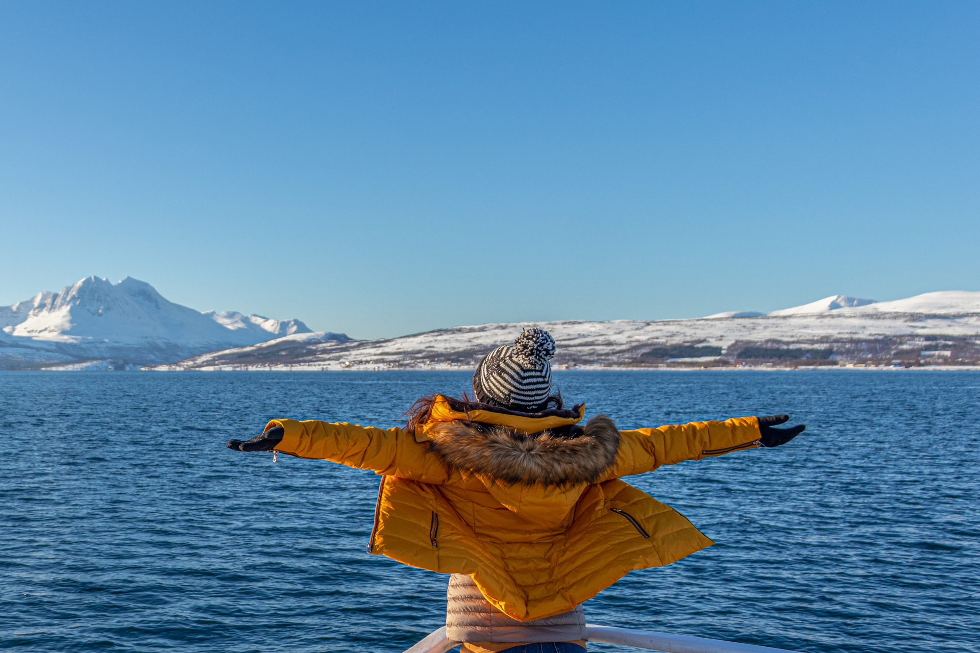 A person in a yellow winter coat and hat stands with outstretched arms on a boat, facing a blue sea and snow-capped mountains under a clear sky.