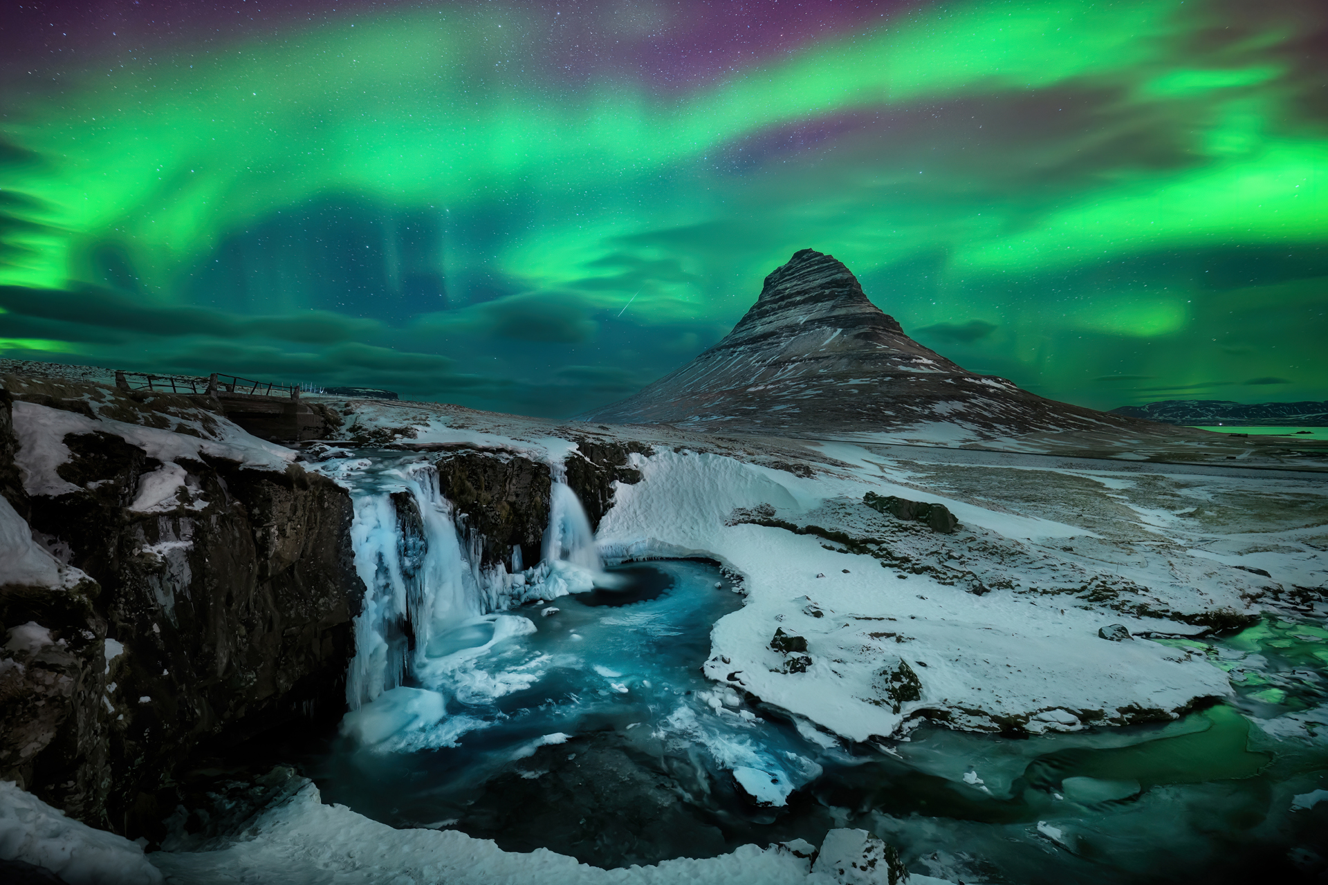 Auroras boreales verdes y moradas surcan el cielo nocturno sobre una montaña nevada, cascadas y un río turquesa.