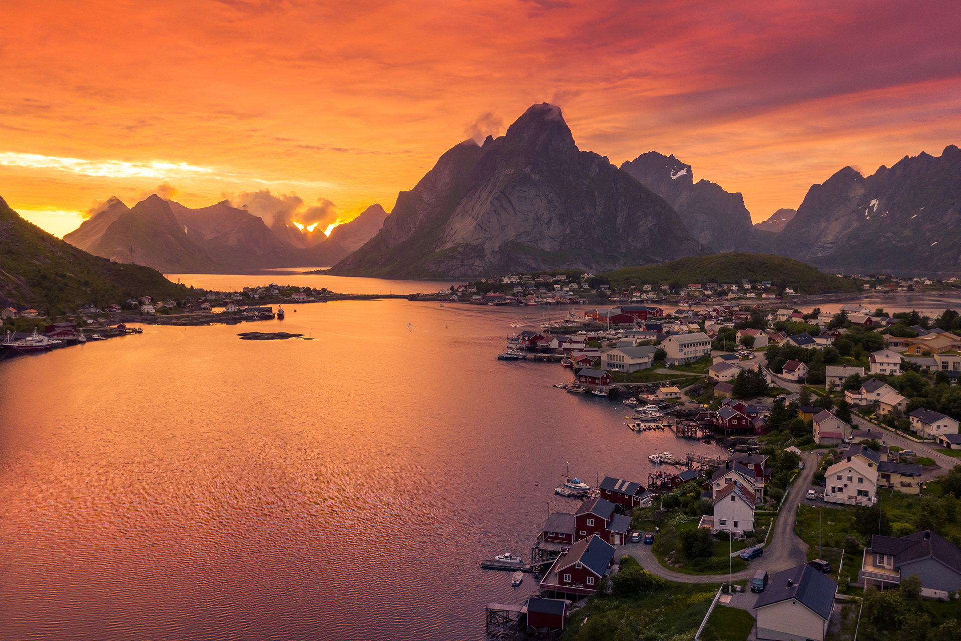A vibrant orange and purple sunset illuminates a coastal village nestled among dramatic, dark mountains and calm waters.