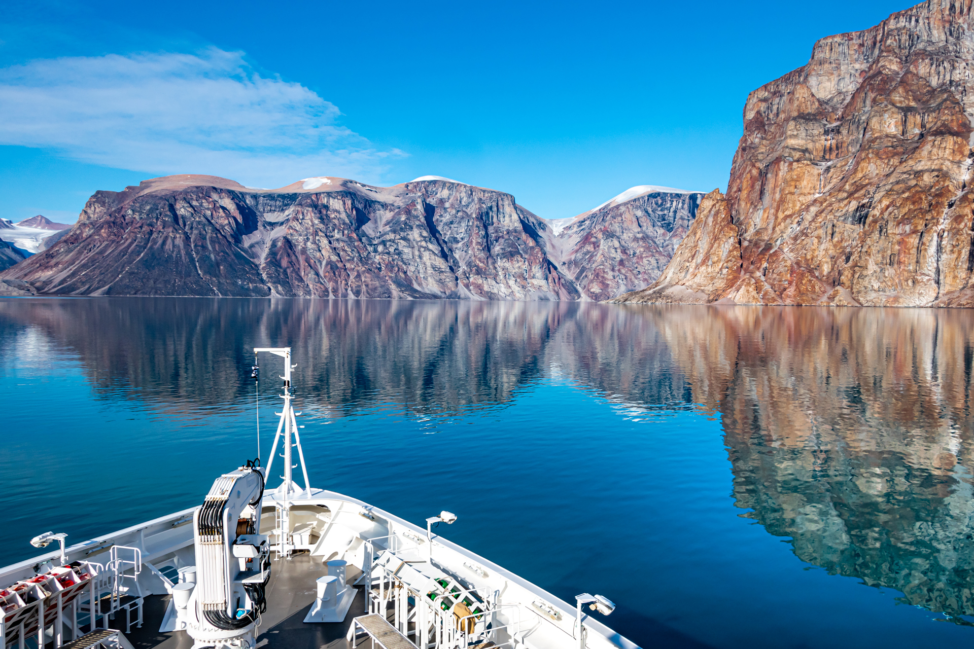 View from the bow of a white ship sailing through calm, deep blue water surrounded by towering, multicolored cliffs under a partly cloudy blue sky.