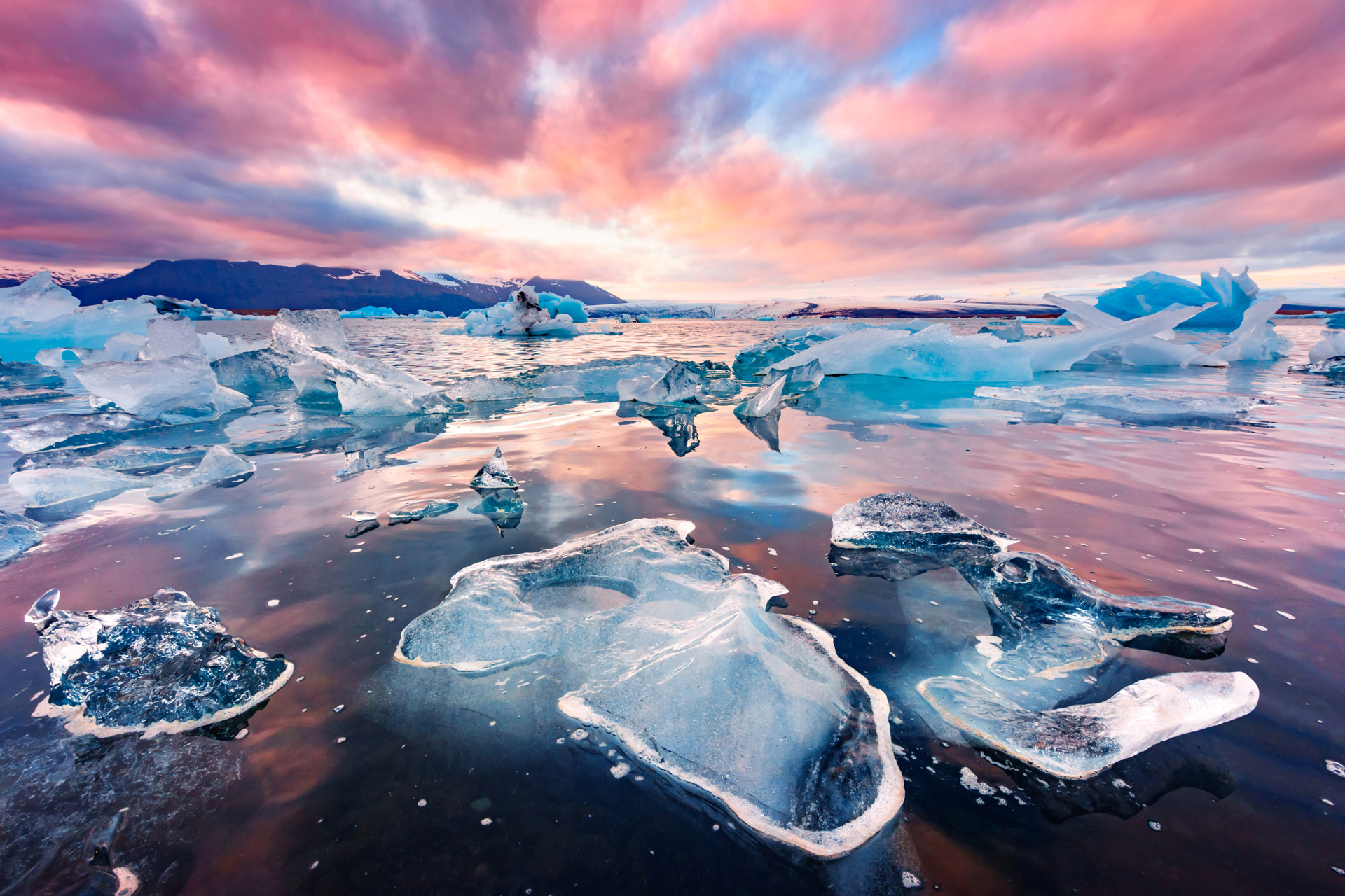 Icebergs and ice floes drift in a calm, reflective body of water under a vibrant, colorful sunset sky.
