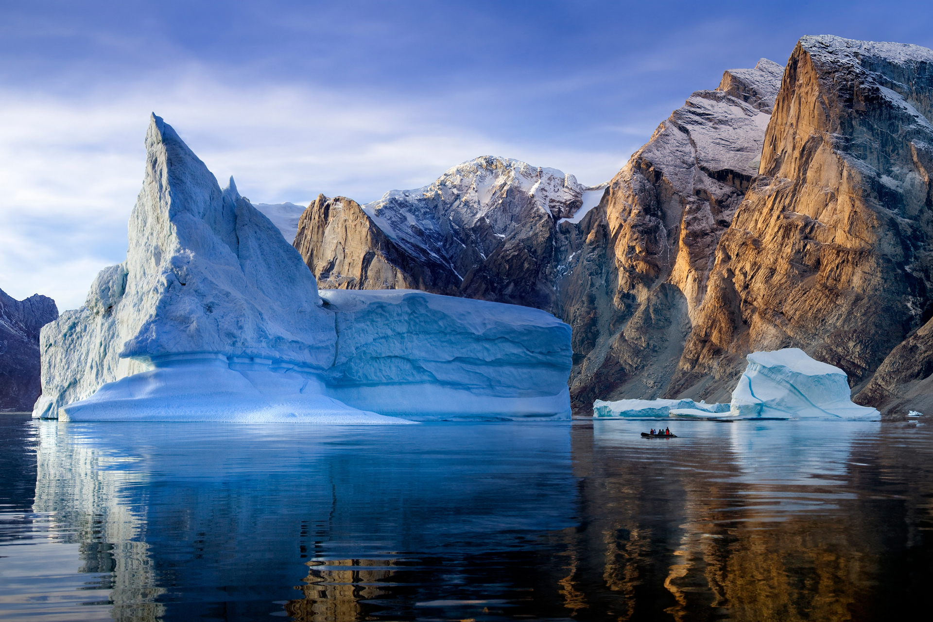 Un gran iceberg azul claro con un pico puntiagudo destaca en un mar azul oscuro en calma, con un pequeño barco rojo y montañas lejanas cubiertas de nieve bajo un cielo claro y brillante.