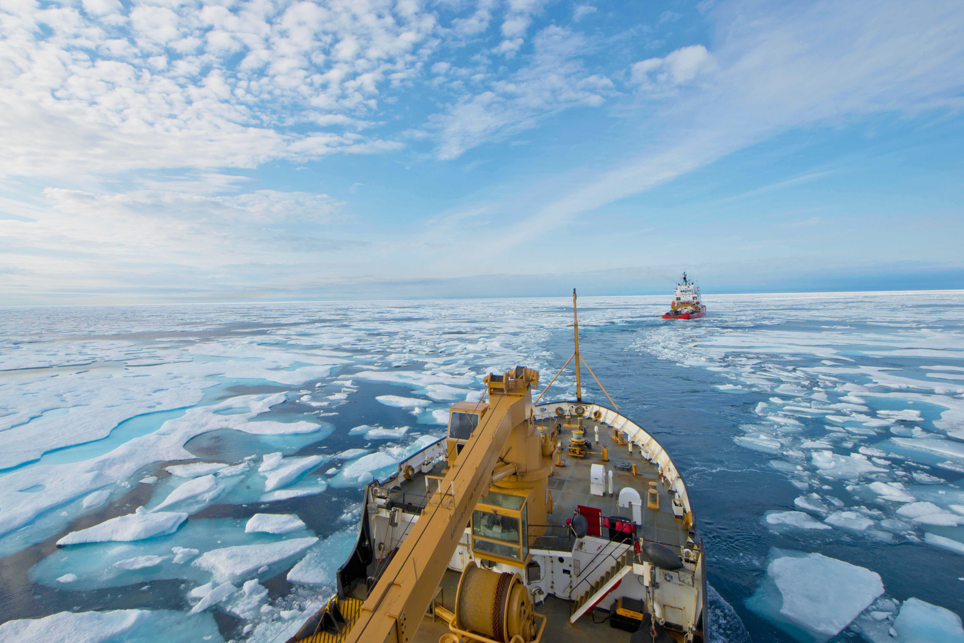 Vista desde la cubierta de un buque rompehielos que navega por un canal a través de una vasta extensión de hielo marino fracturado bajo un cielo parcialmente nublado, con otro rompehielos visible en la distancia.