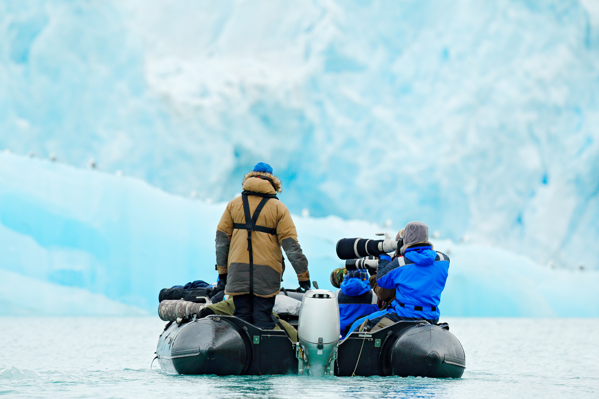 Dos personas en una lancha neumática negra sobre aguas agitadas, una de pie y otra de rodillas con una gran cámara y un objetivo, frente a un gran glaciar azul claro.