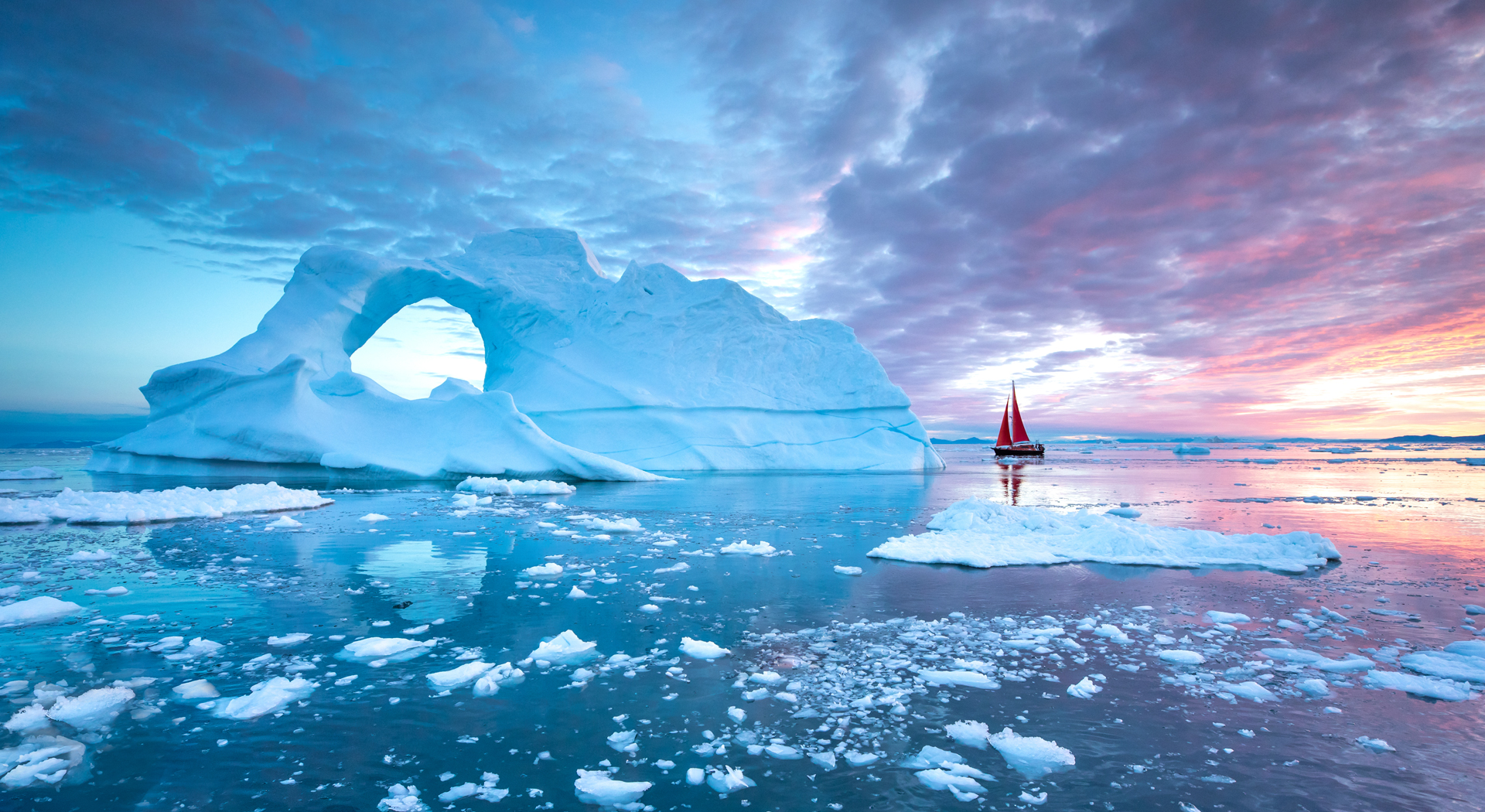 A large, arched iceberg dominates a cold, choppy sea dotted with smaller ice chunks, with a red-sailed boat in the distance under a cloudy sky with hues of pink and blue.